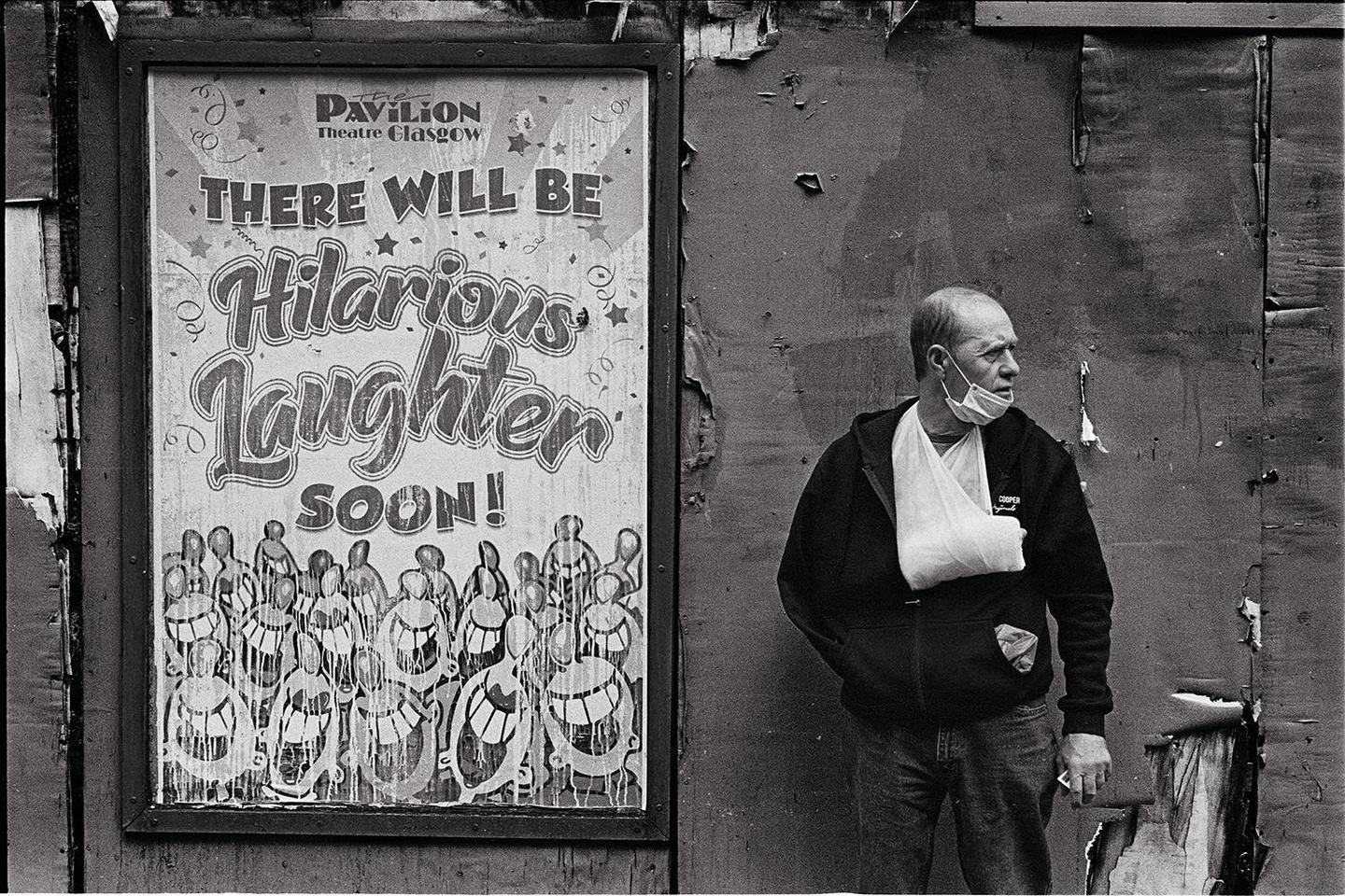 A man with a face mask and arm sling stands next to a poster advertising "Hilarious Laughter Soon!" in Glasgow.