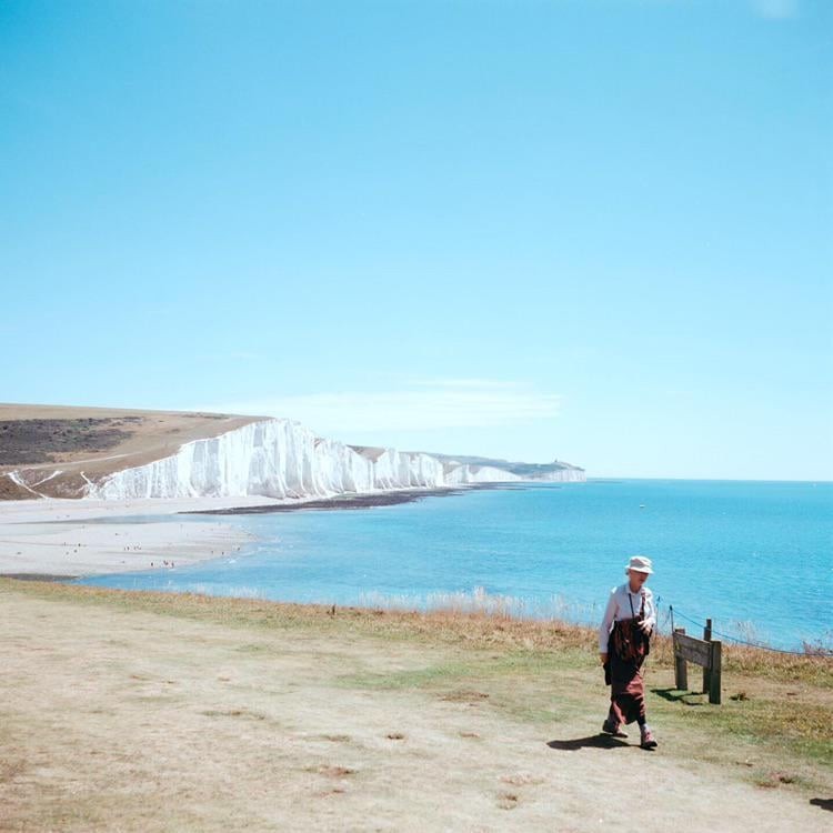 A woman walks along a grassy cliff overlooking a beach and white cliffs next to a bright blue sea.
