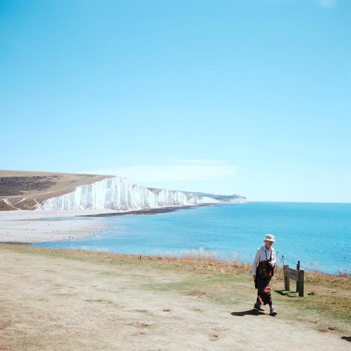 A woman walks along a grassy cliff overlooking a beach and white cliffs next to a bright blue sea.