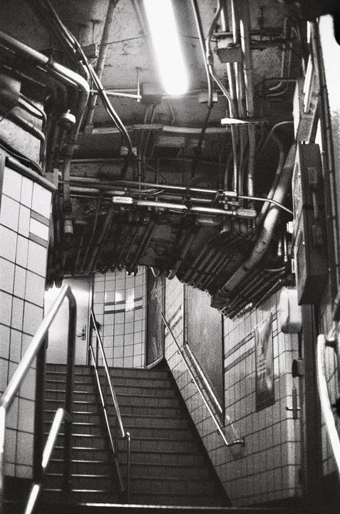 A black and white photograph shows a subway staircase with pipes and wires on the ceiling.