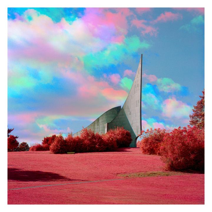 A concrete building with a sharp, pointed roof stands in a field of red grass under a sky filled with colorful clouds.