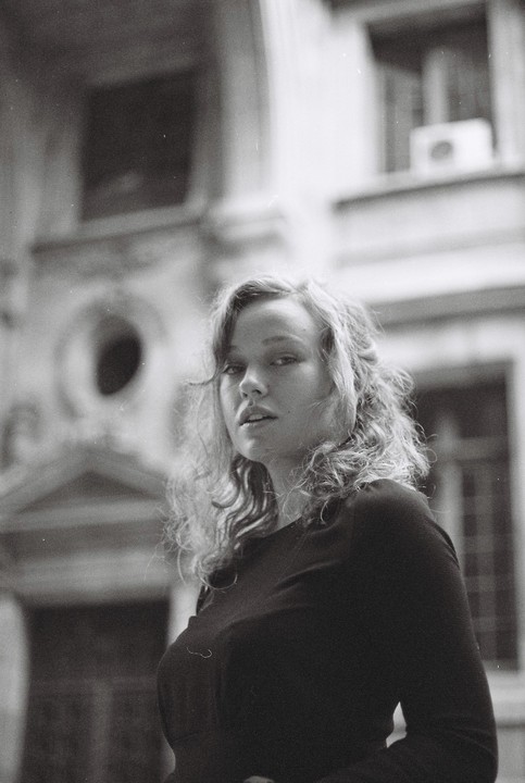 A woman with curly hair looks at the viewer, with a building in the background.