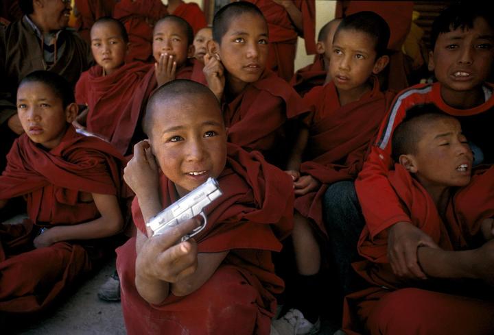 A boy in a red robe holds a toy gun while sitting among other boys in red robes.