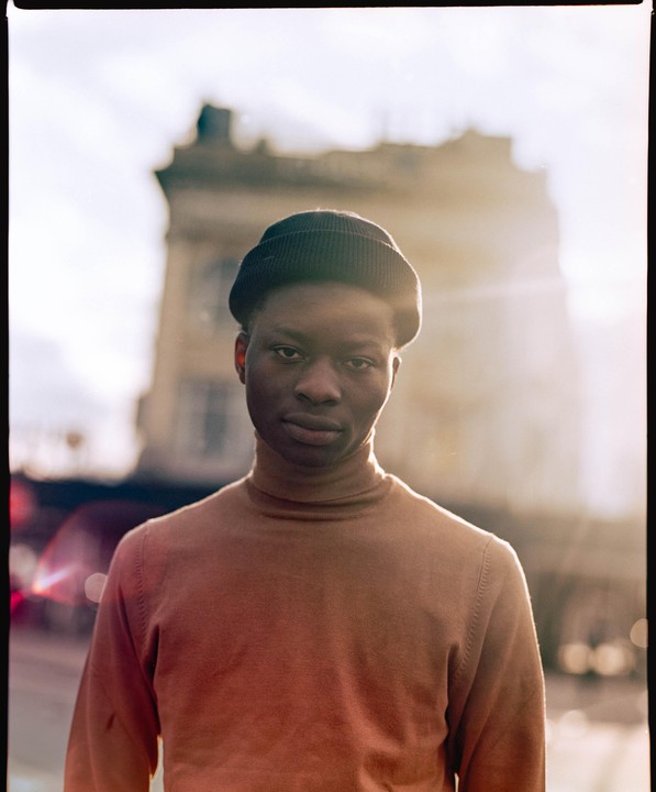 A man in a beanie and turtleneck stands in front of a building with bright light behind him.