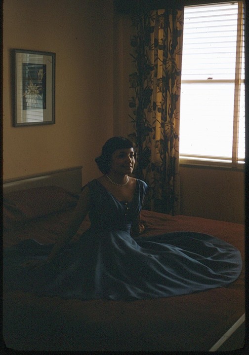 A woman in a blue dress and pearl necklace sits on a bed next to a window with blinds and a patterned curtain.