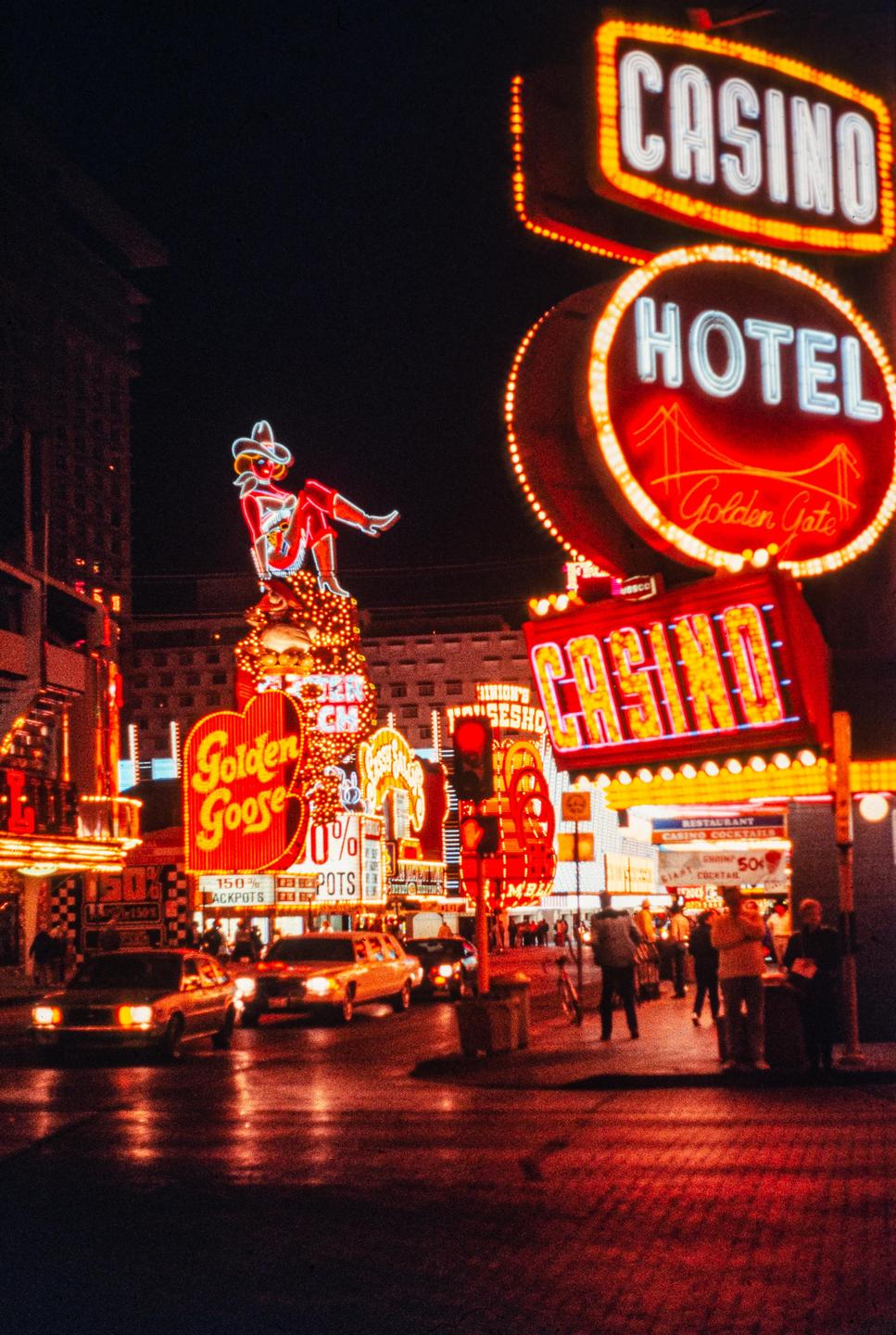 A street at night is illuminated by numerous neon signs for casinos and hotels, with cars driving on the wet road and people walking on the sidewalk.