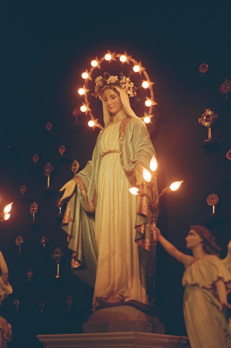 A statue of a woman with a halo of stars and flowers on her head stands in a dark room, with another statue of an angel holding a candelabra visible to the right.