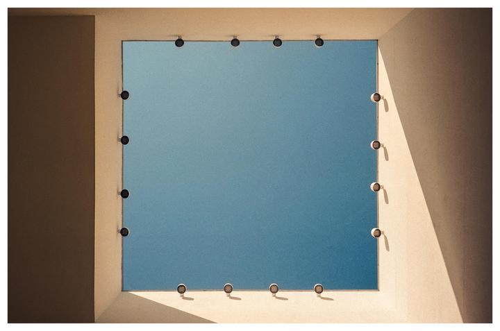 A square opening in a building frames a clear blue sky, with small lights lining the edges of the opening.