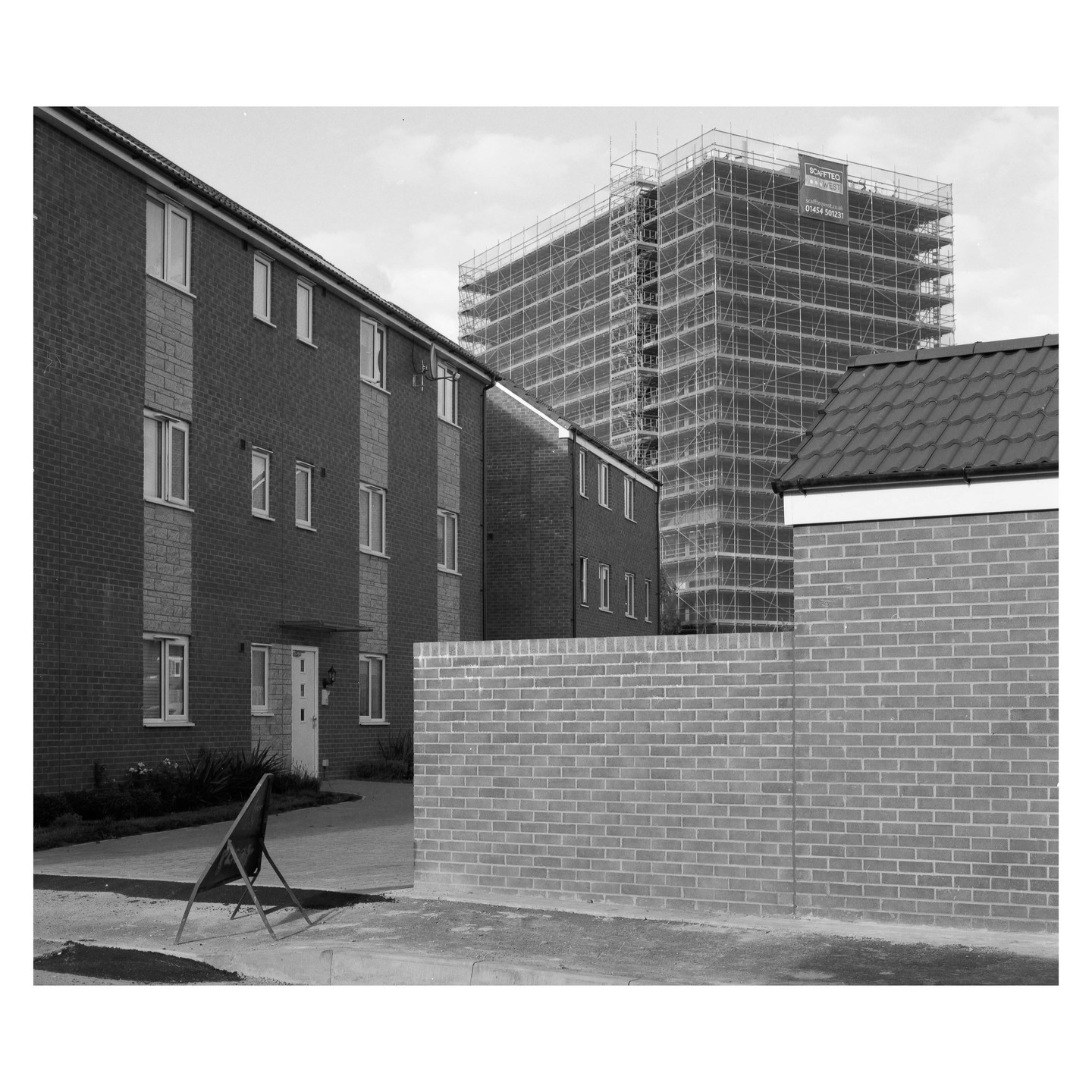 A black and white image shows a tall building covered in scaffolding behind a brick wall and residential buildings.