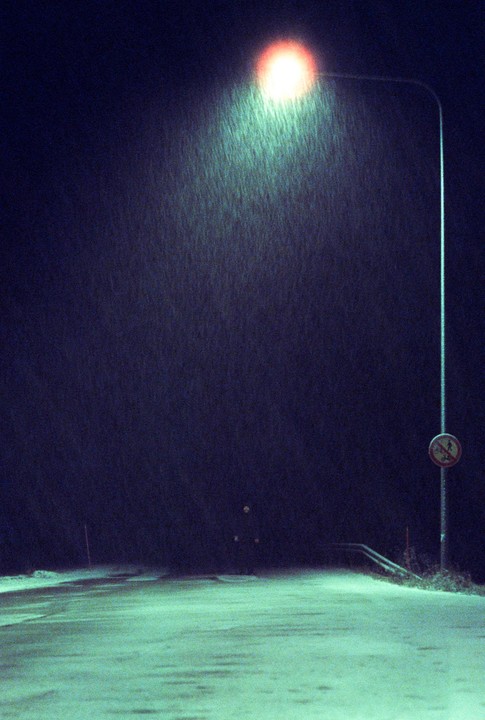 A street light illuminates falling snow on a dark night, with a street sign visible on the right.