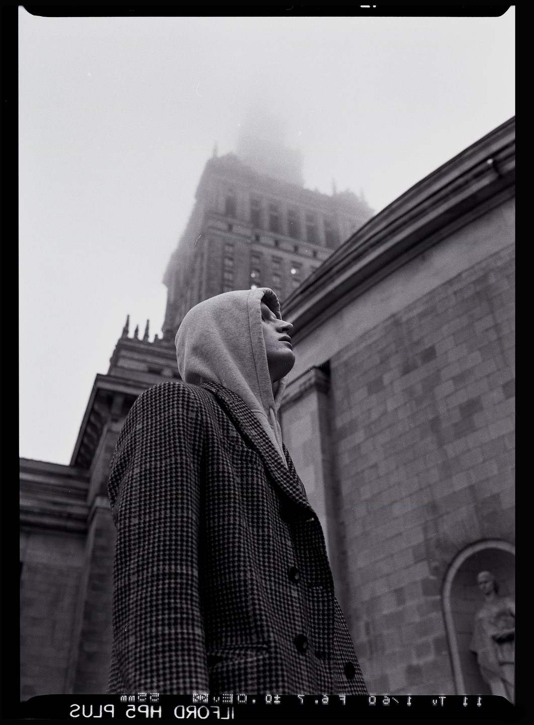 A man in a hooded sweatshirt and patterned coat looks up at a tall building partially obscured by fog.