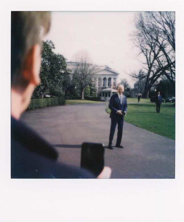 A man in a suit stands on a paved path in front of a large white building with columns, while another man in the foreground holds a phone.