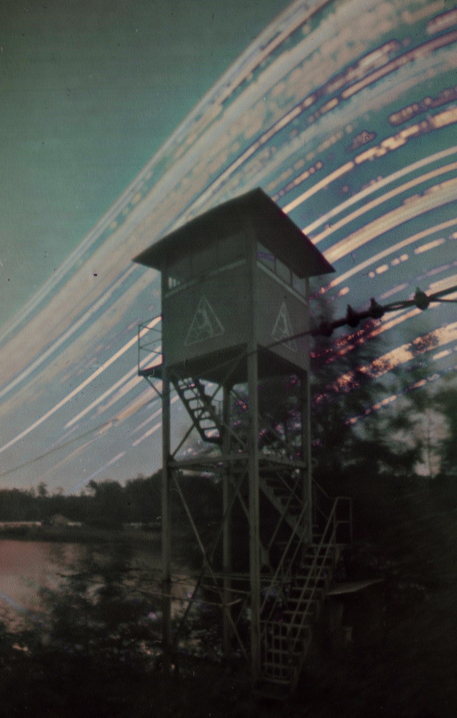 A watchtower stands by a lake under a sky with long light trails.
