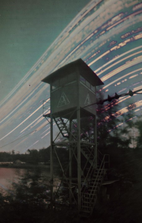 A watchtower stands by a lake under a sky with long light trails.