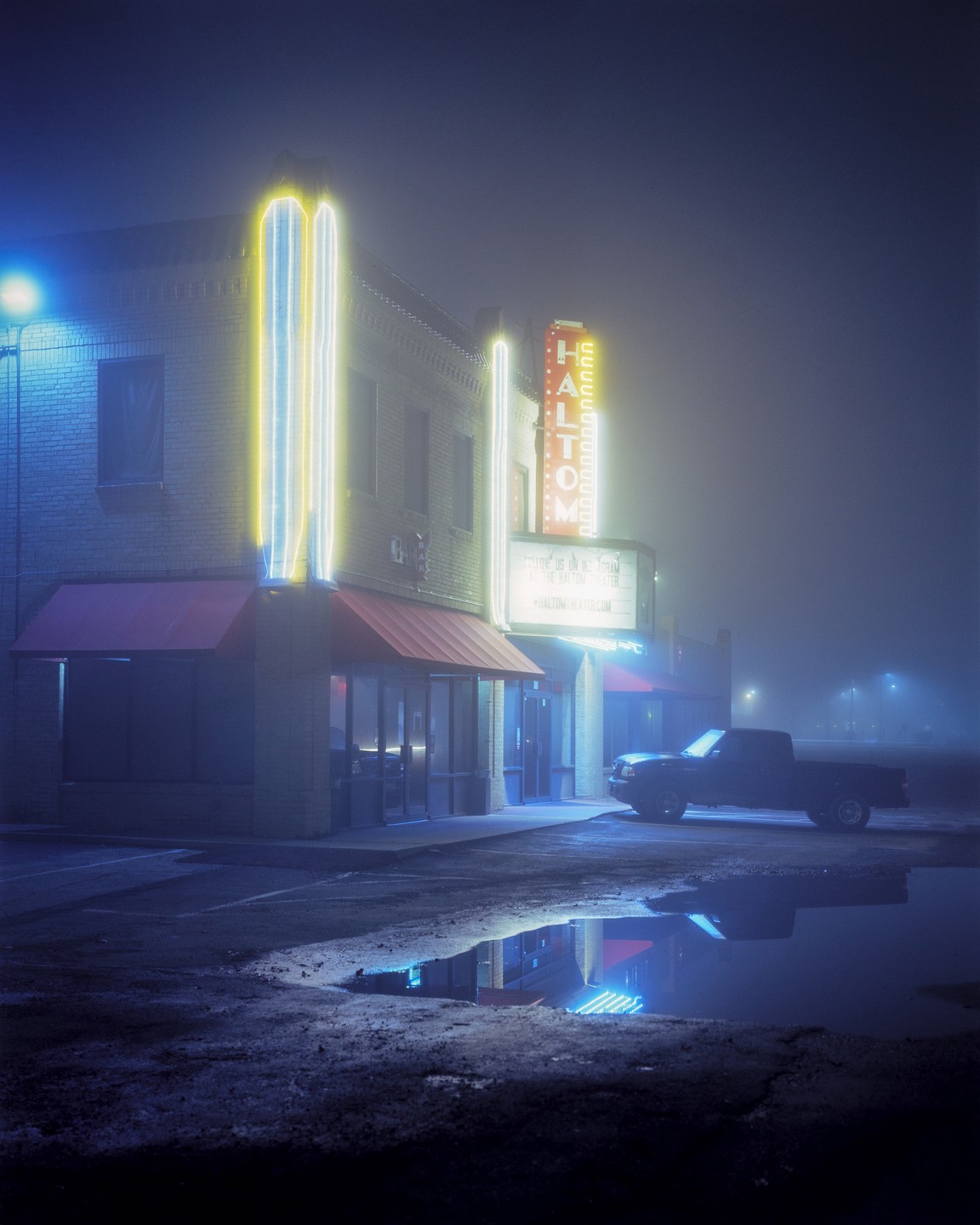 A neon-lit building with a red awning and a truck parked in front is reflected in a puddle on a foggy night.