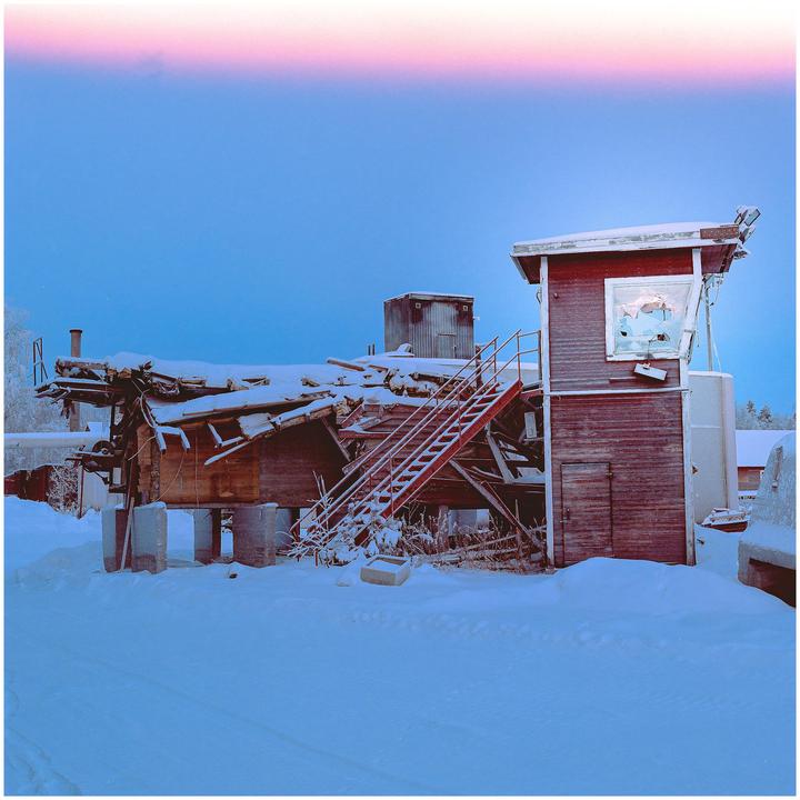 A snow-covered abandoned rail yard with a partially collapsed building and a small office building with a broken window is visible under a blue sky with a pink light leak.