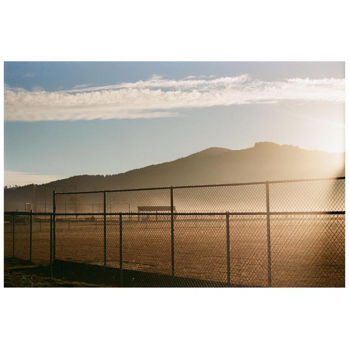 A chain link fence runs across the foreground of a hazy field with a mountain in the background under a blue sky with clouds.