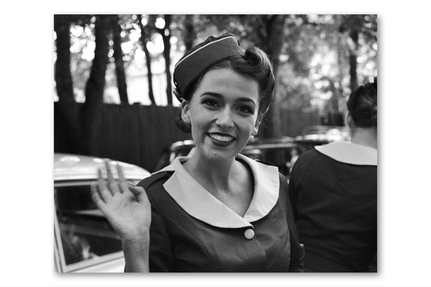 A woman in a uniform with a hat smiles and waves at the camera.