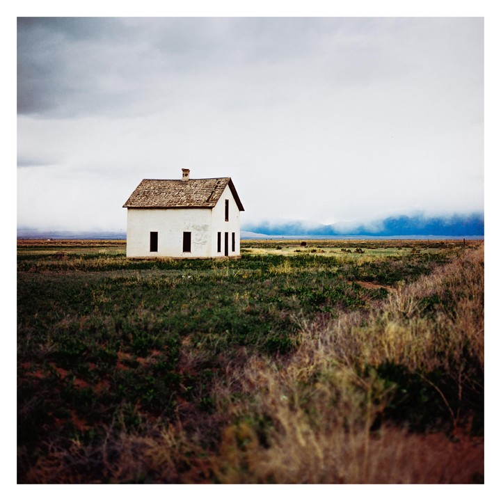 A small white house with a brown roof stands alone in a vast green field under a cloudy sky.