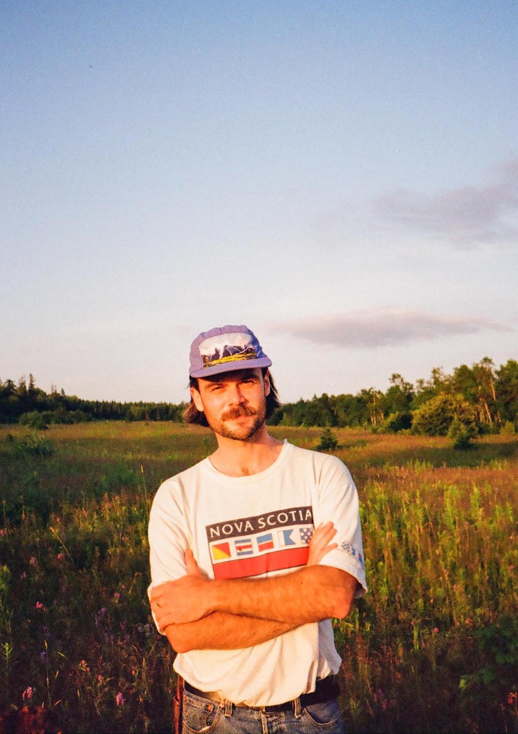 A man with a mustache and a hat stands in a field with his arms crossed.