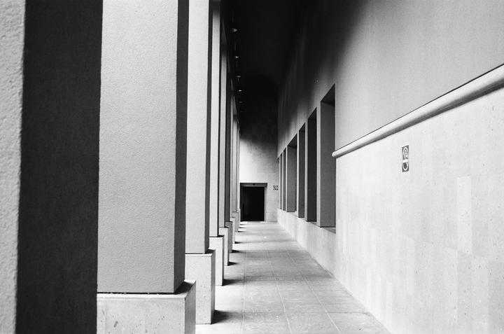 A long, narrow hallway with a row of pillars on the left and a row of windows on the right leads to a dark doorway.