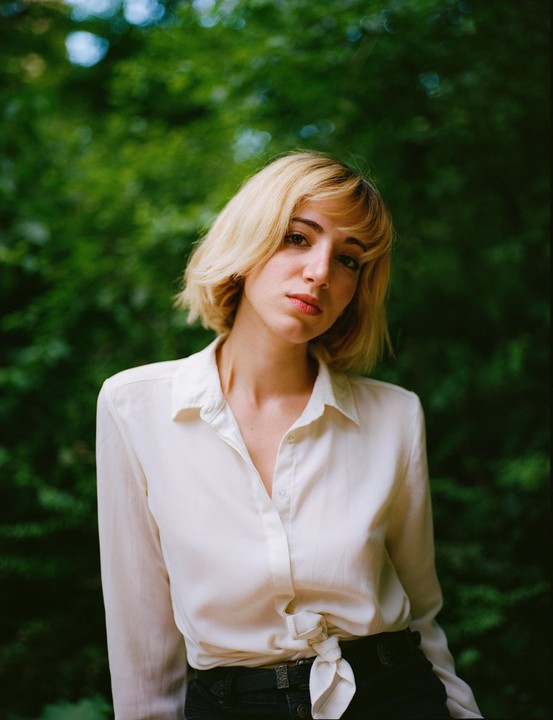 A woman with blonde hair and a white shirt stands in front of green foliage.