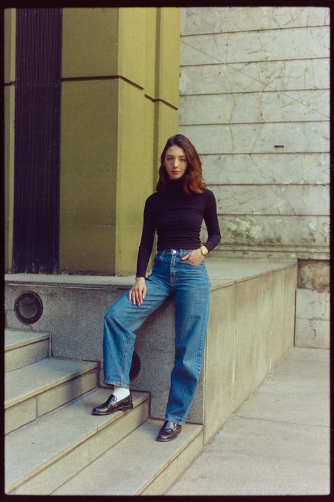 A woman wearing a black turtleneck and jeans stands on concrete steps next to a building.