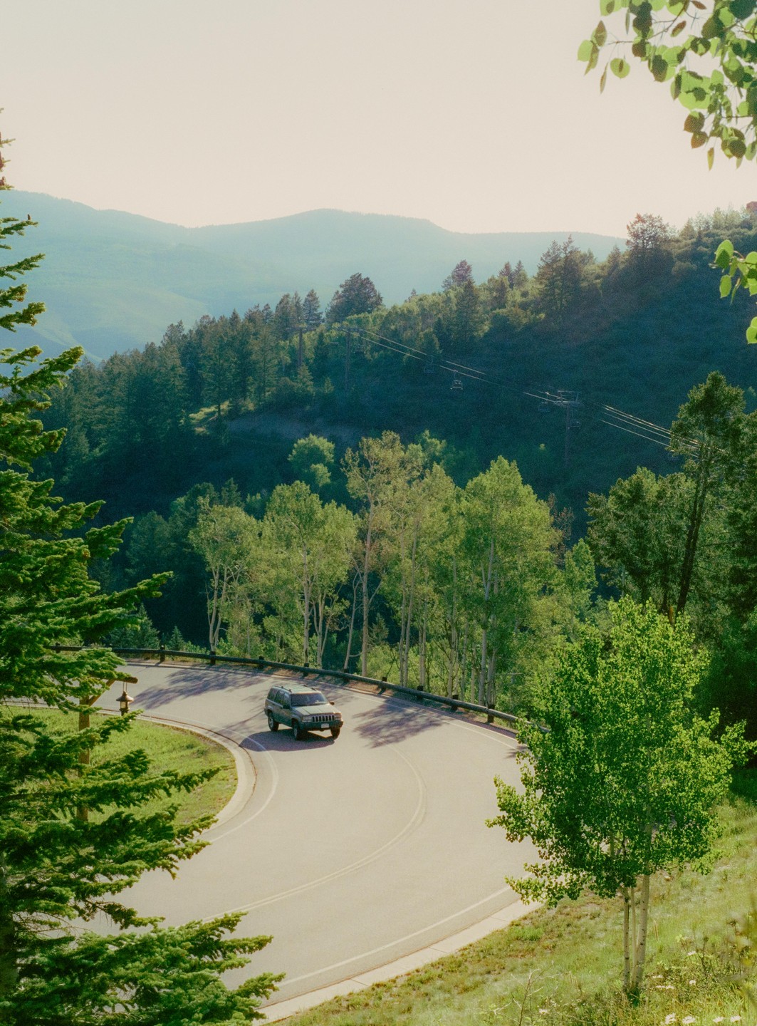 A car drives on a winding mountain road surrounded by lush green trees and distant hills under a bright sky.