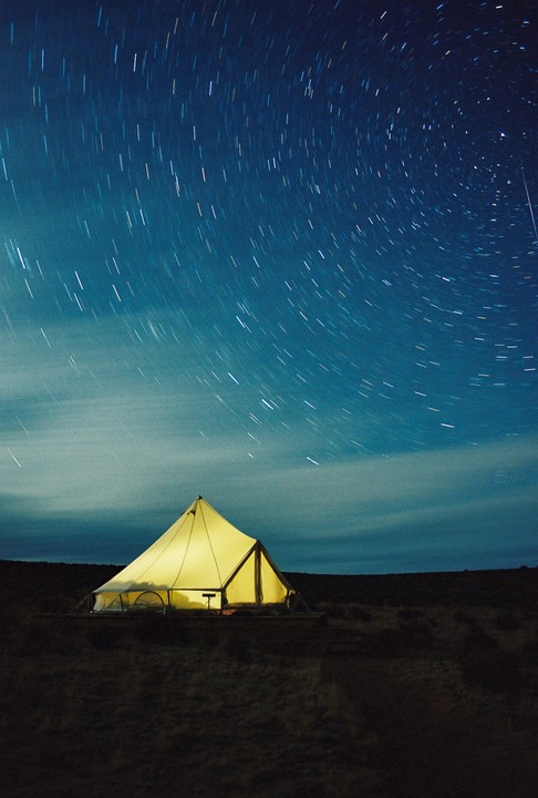 A glowing tent sits under a night sky filled with star trails.
