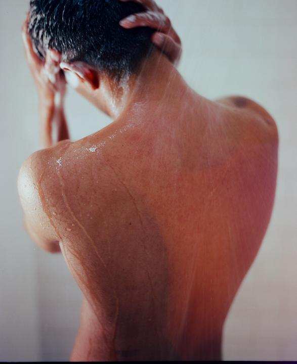 A man is showering, seen from behind, with water and shampoo running down his back and hair.