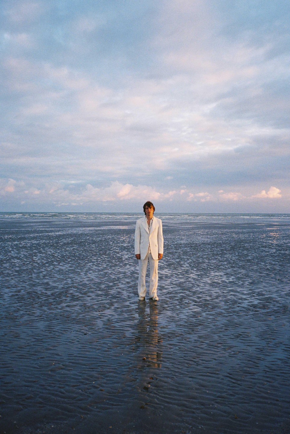 A man in a white suit stands on a wet beach at dusk with a cloudy sky above.