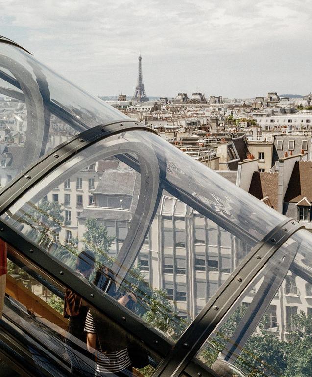 A woman rides an escalator with a view of the Eiffel Tower and the city of Paris in the background.
