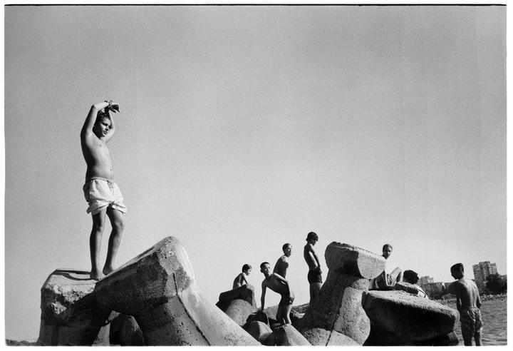 A group of boys stands on concrete tetrapods by the water under a clear sky.