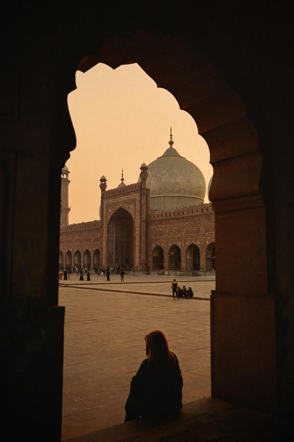 A woman sits in the foreground looking out at a large mosque with a dome and minarets, framed by an archway.