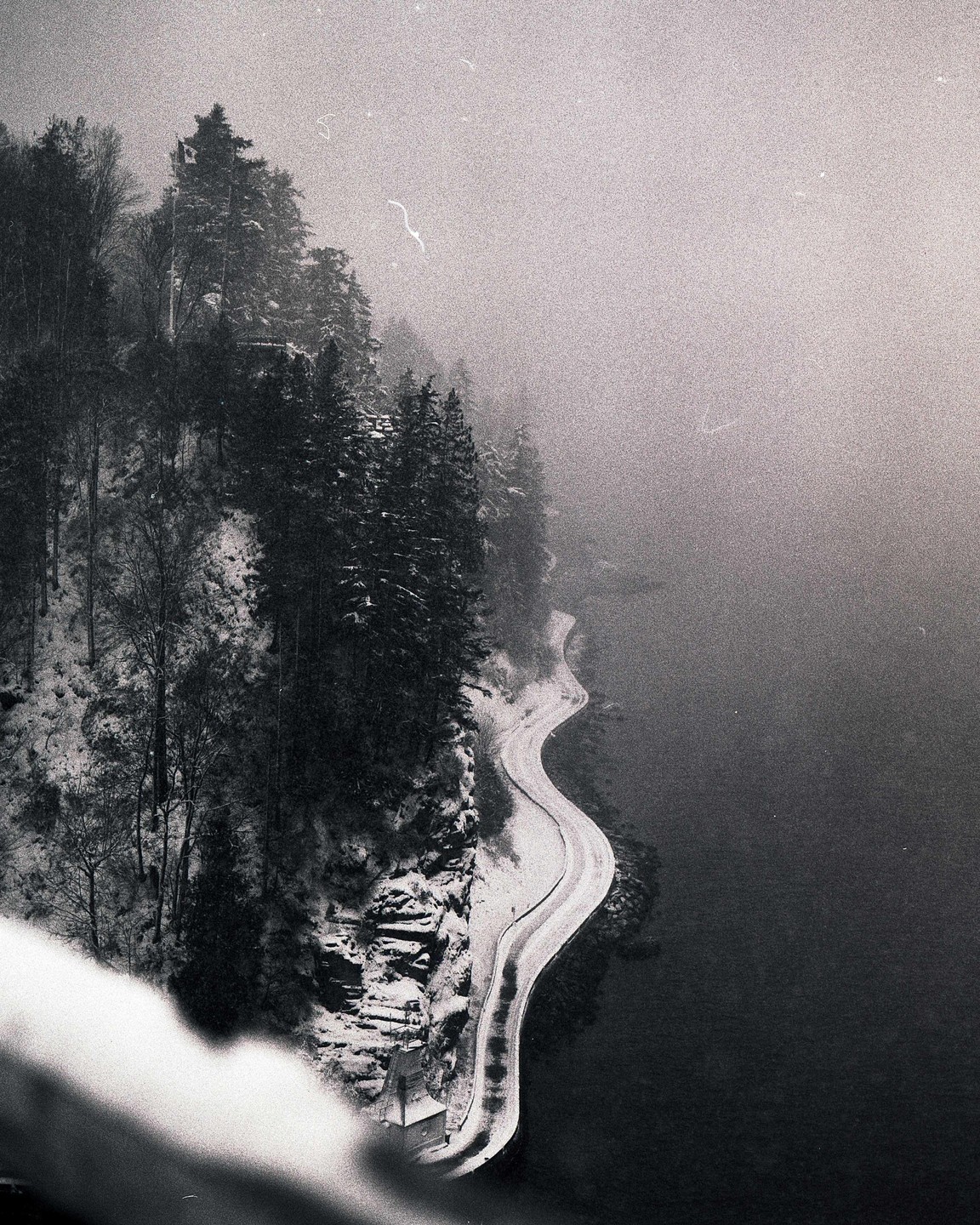 A winding road covered in snow runs alongside a body of water, with snow-covered trees on a steep bank under a hazy sky.