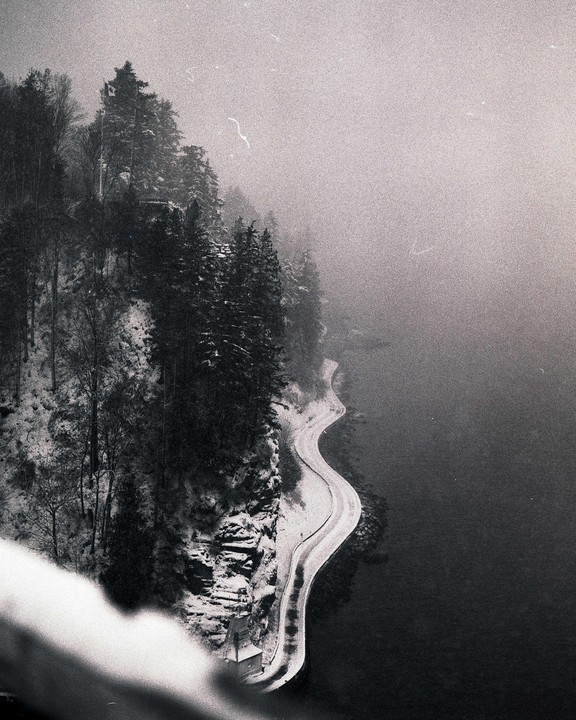 A winding road covered in snow runs alongside a body of water, with snow-covered trees on a steep bank under a hazy sky.
