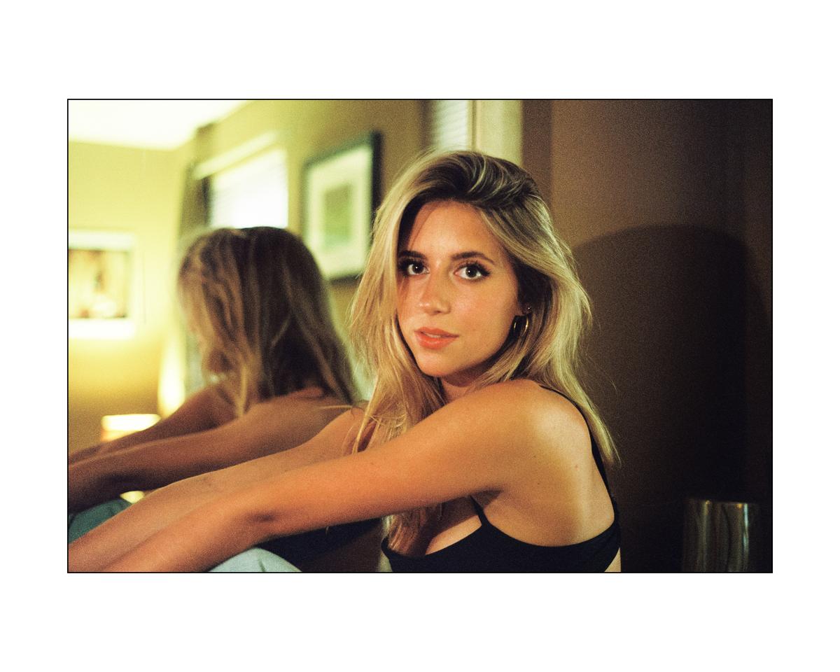 A woman with long blonde hair looks at the viewer while her reflection is visible in a mirror behind her.
