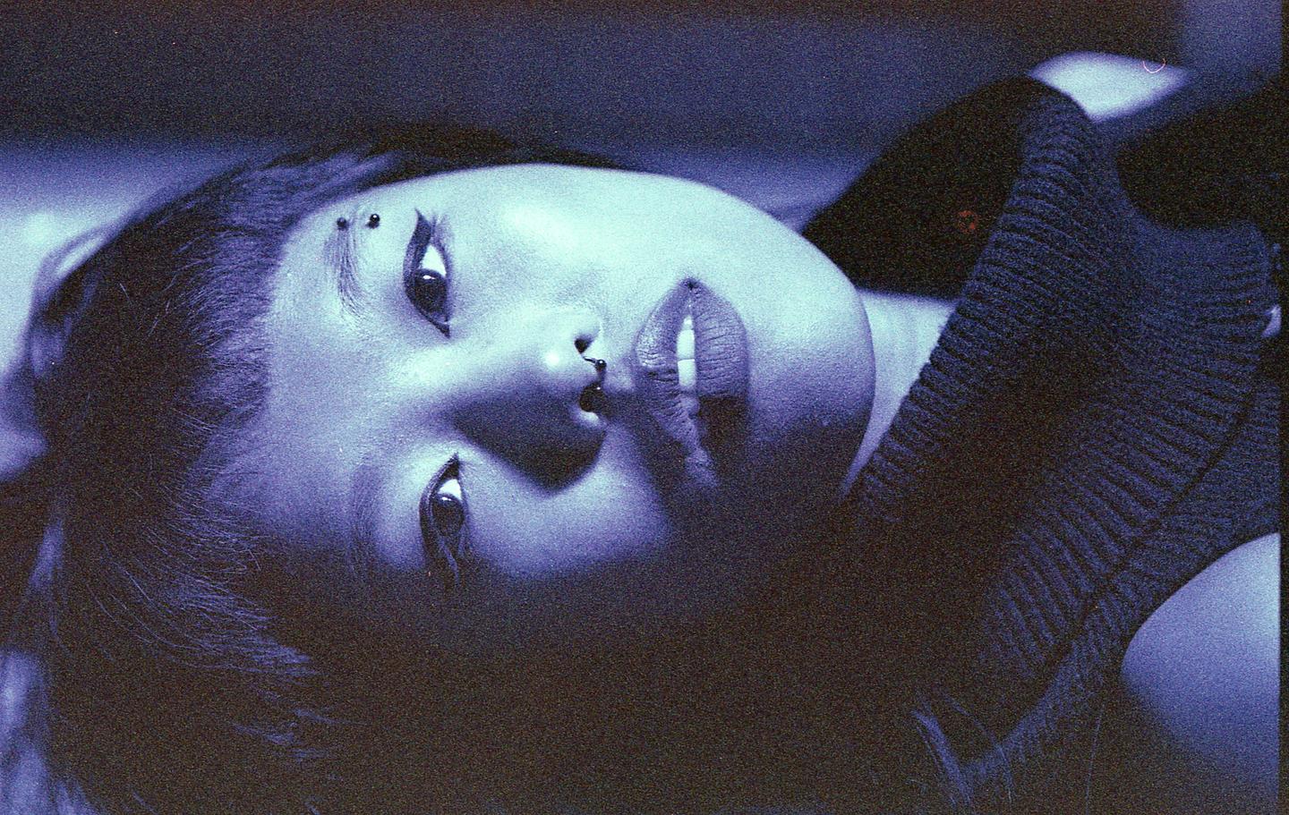 A woman with dark hair and facial piercings looks up at the viewer.