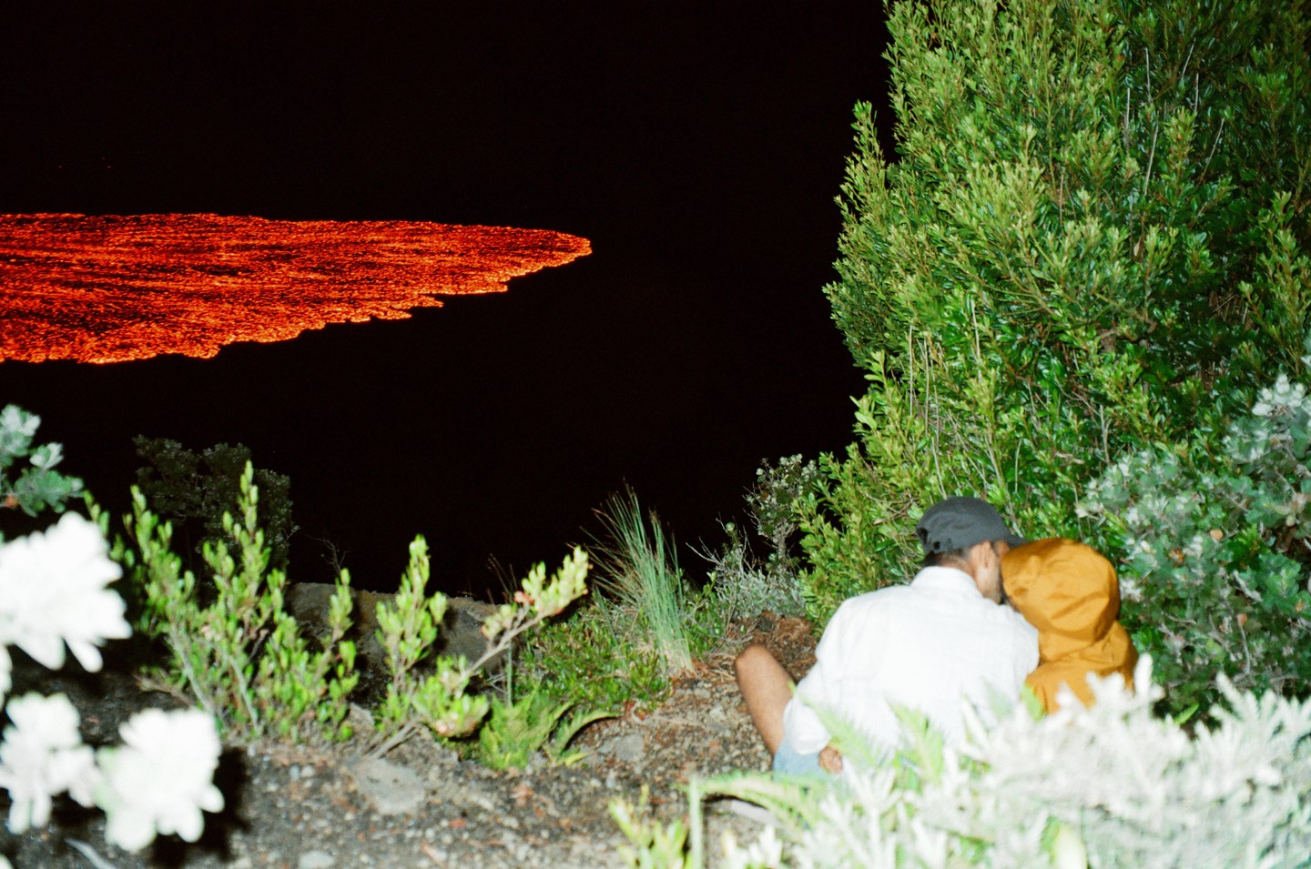 A couple sits in foliage at night, looking at a glowing red lava flow.