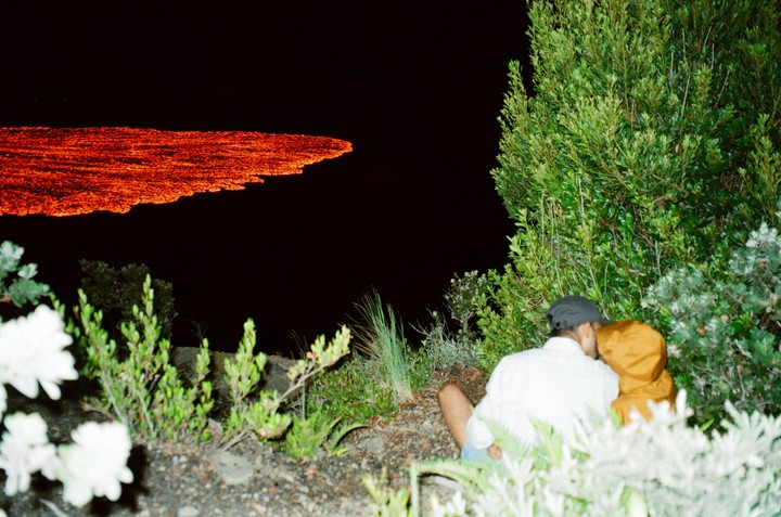 A couple sits in foliage at night, looking at a glowing red lava flow.