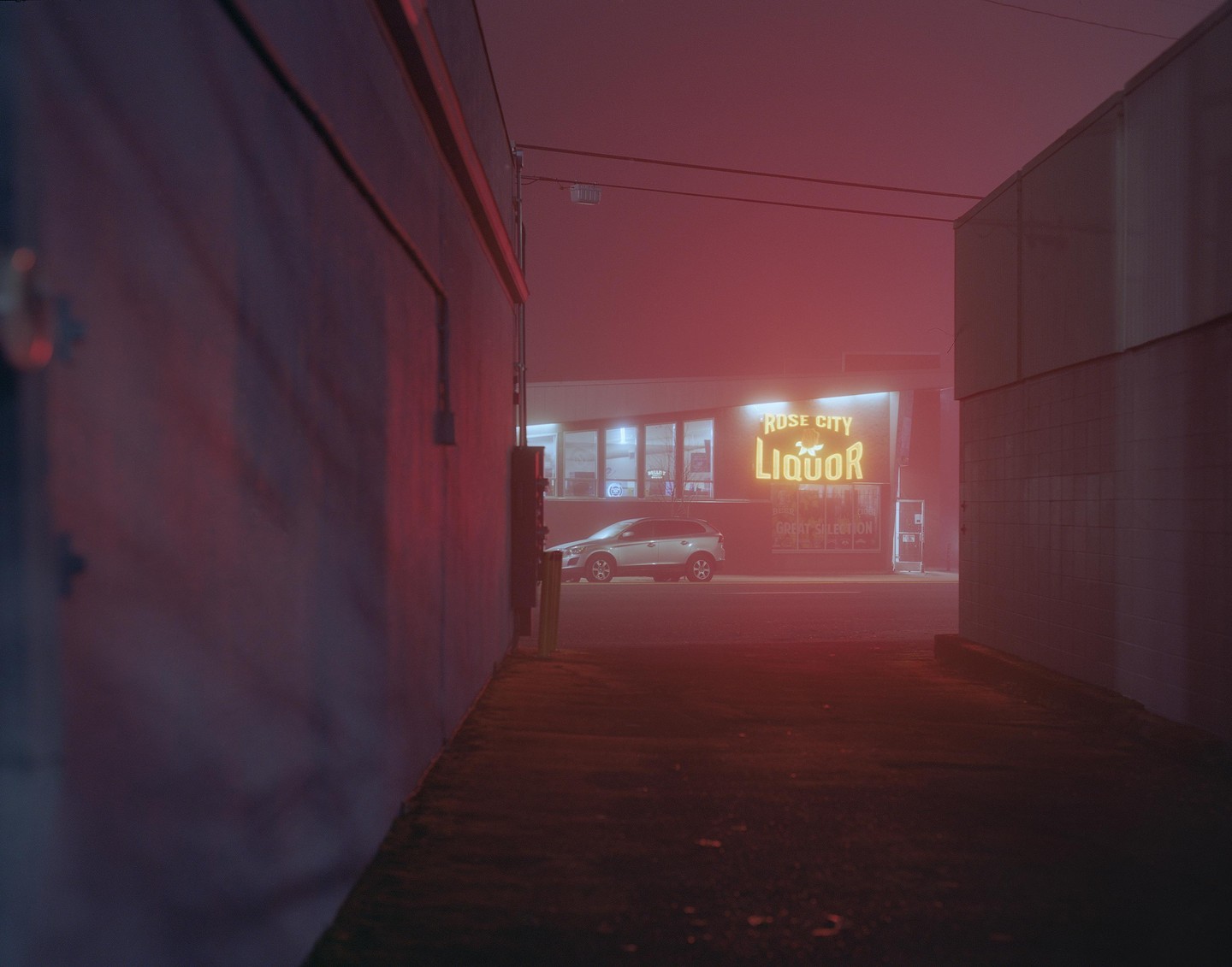 A silver car is parked in front of a liquor store with a neon sign that reads "Rose City Liquor" on a foggy night.