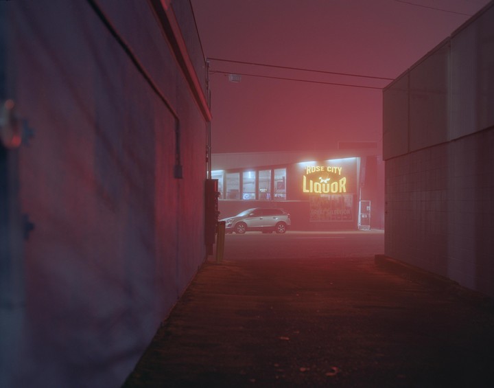 A silver car is parked in front of a liquor store with a neon sign that reads "Rose City Liquor" on a foggy night.