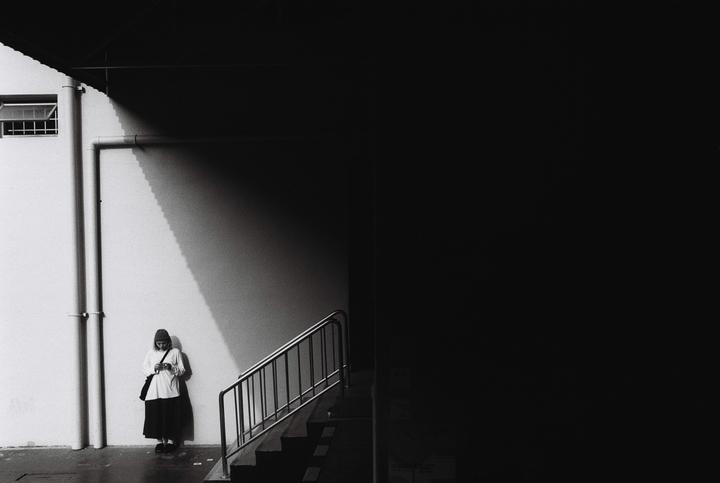 A woman stands against a wall with a strong shadow line next to a staircase.