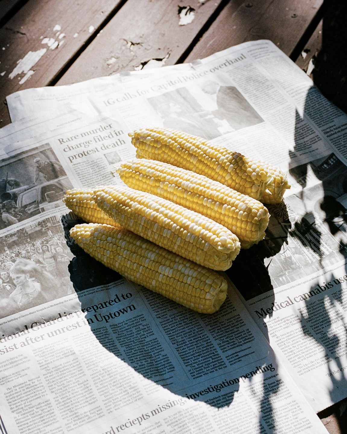 Four ears of corn rest on a newspaper on a wooden surface, with shadows cast across the scene.