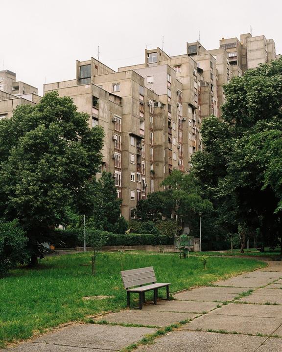 A concrete apartment building rises behind a park bench and a path.