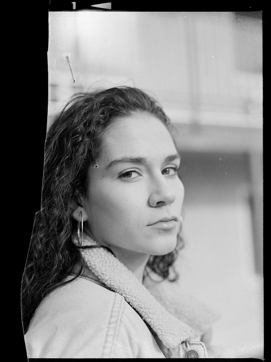 A woman with curly hair and a denim jacket with a sherpa collar looks directly at the viewer.