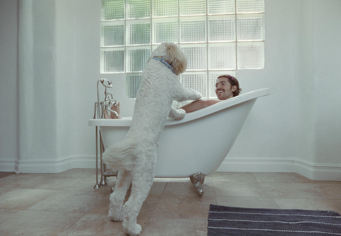 A white dog stands on its hind legs with its paws on the edge of a bathtub, looking at a man who is smiling and relaxing in the tub.