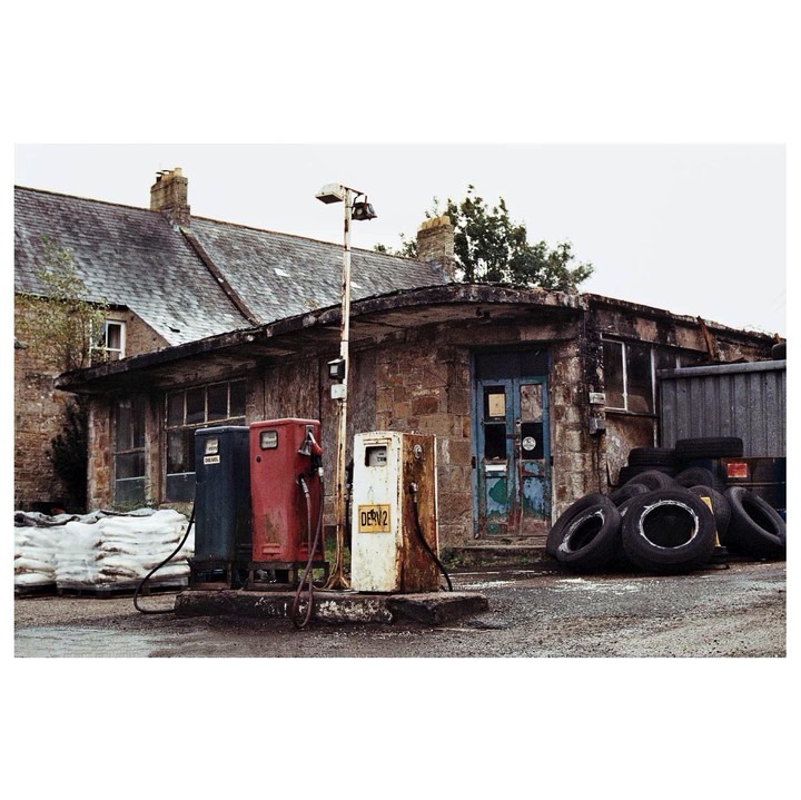 Three old fuel pumps stand in front of a stone building with a blue door and a pile of tires.