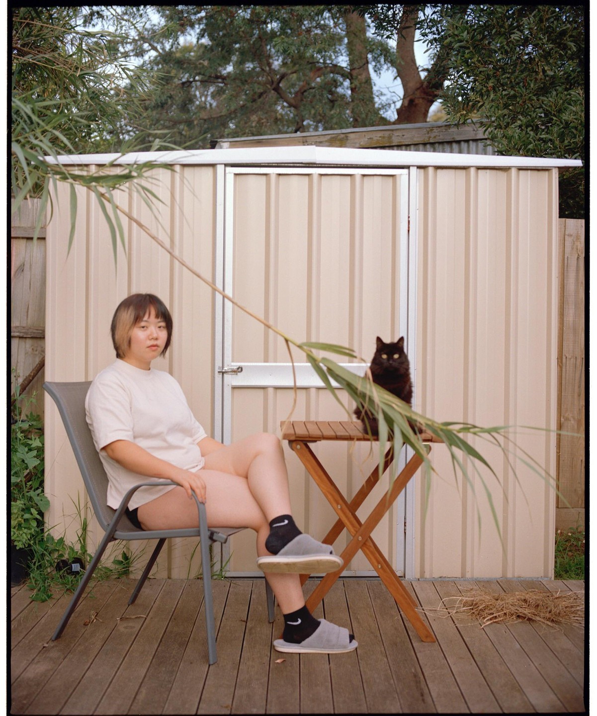 A woman sits on a chair next to a small table with a black cat on it, in a backyard.