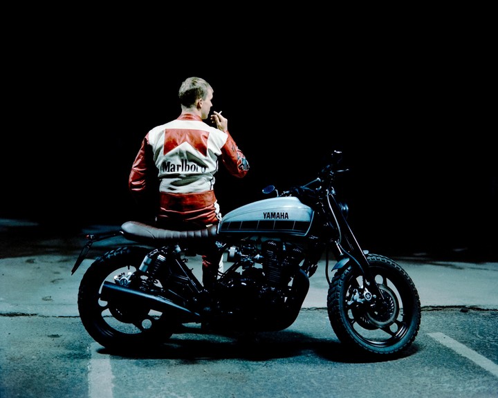 A man in a racing jacket sits on a motorcycle and smokes a cigarette at night.